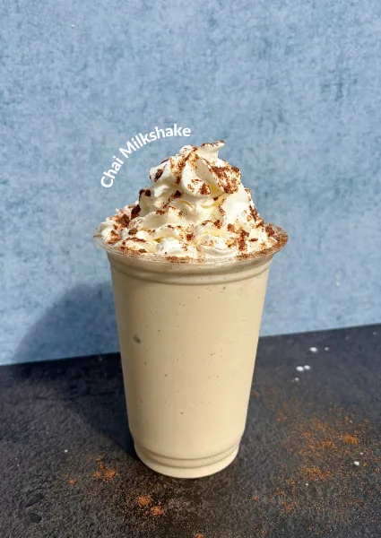 A chai milkshake in a clear cup piled with whipped cream and dusted with cinnamon, on a dark slate