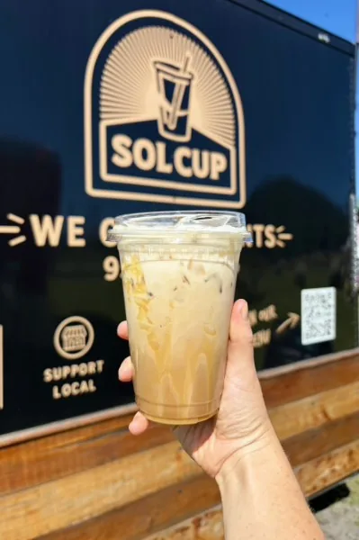 A hand holding an iced latte in a clear cup in front of the black Sol Cup trailer