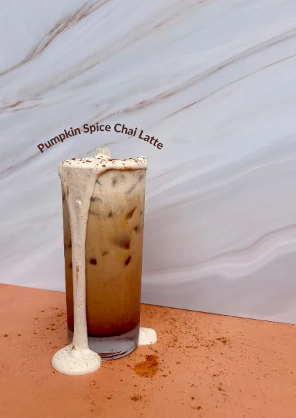 A tall iced pumpkin spice chai latte with cream running down the glass, on a terracotta surface