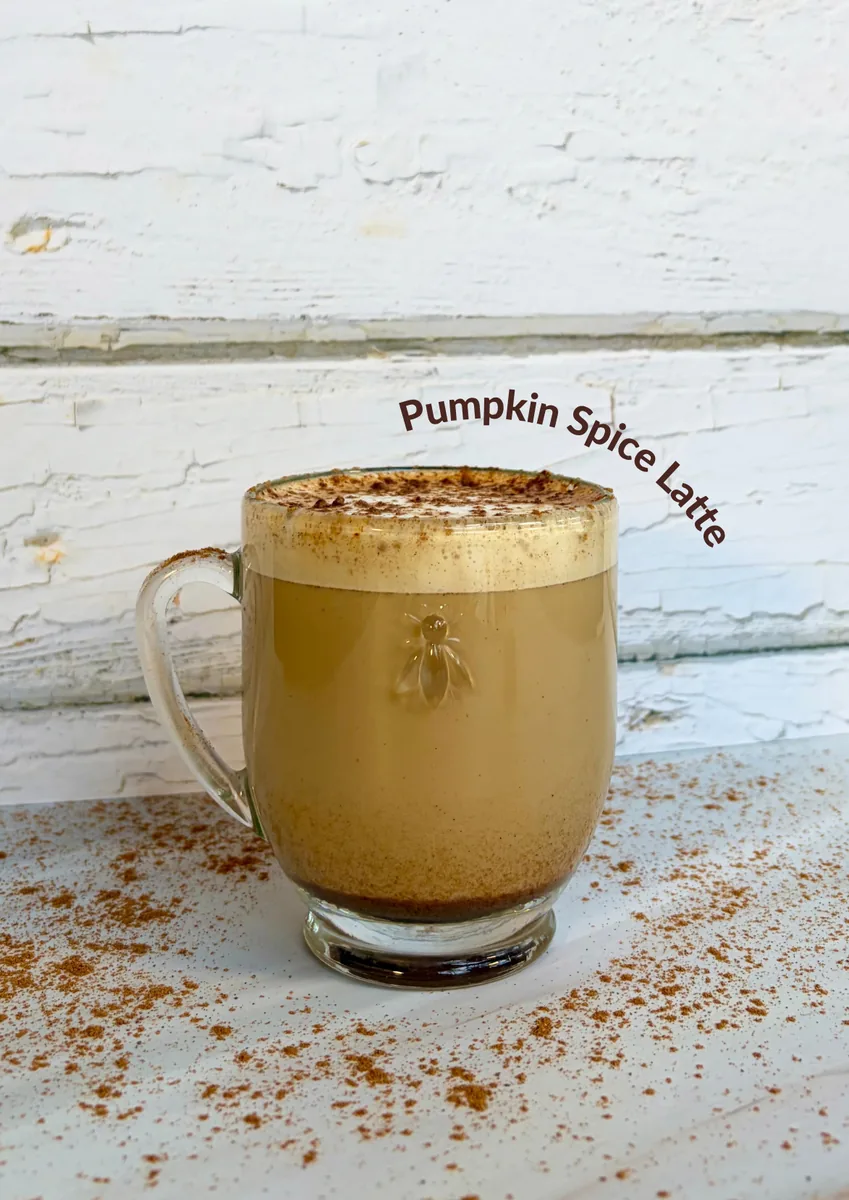 A glass mug of pumpkin spice latte with a thick foam top dusted with cinnamon, on a white table scattered with cinnamon